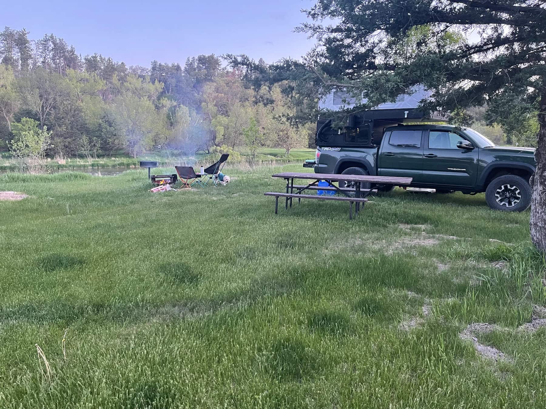 Truck camper parked at Nebraska Falls campsite beside smoldering fire and picnic table. 