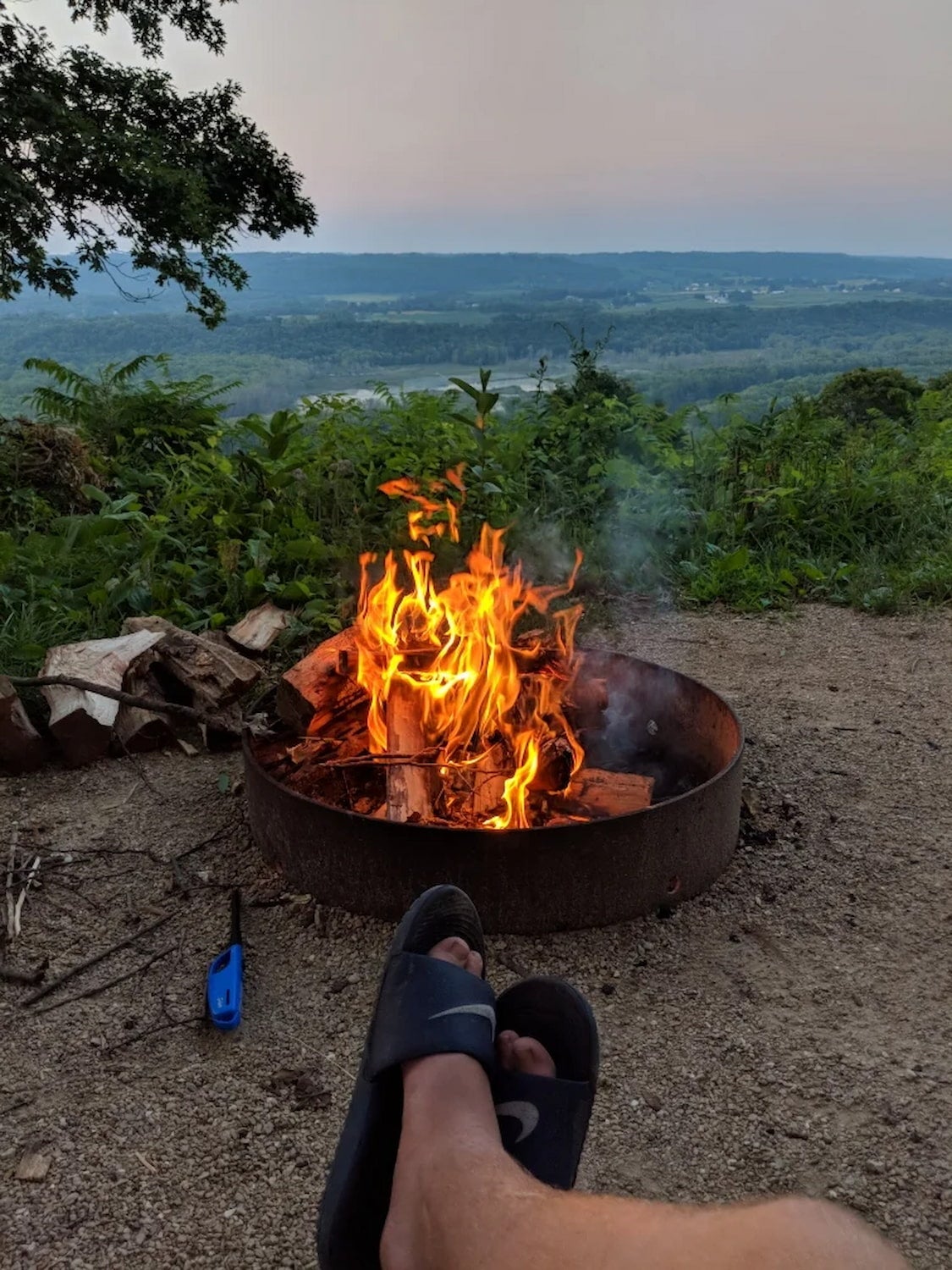 Point of view image showing legs of the photographer crossed i front of fire pit overlooking valley below.