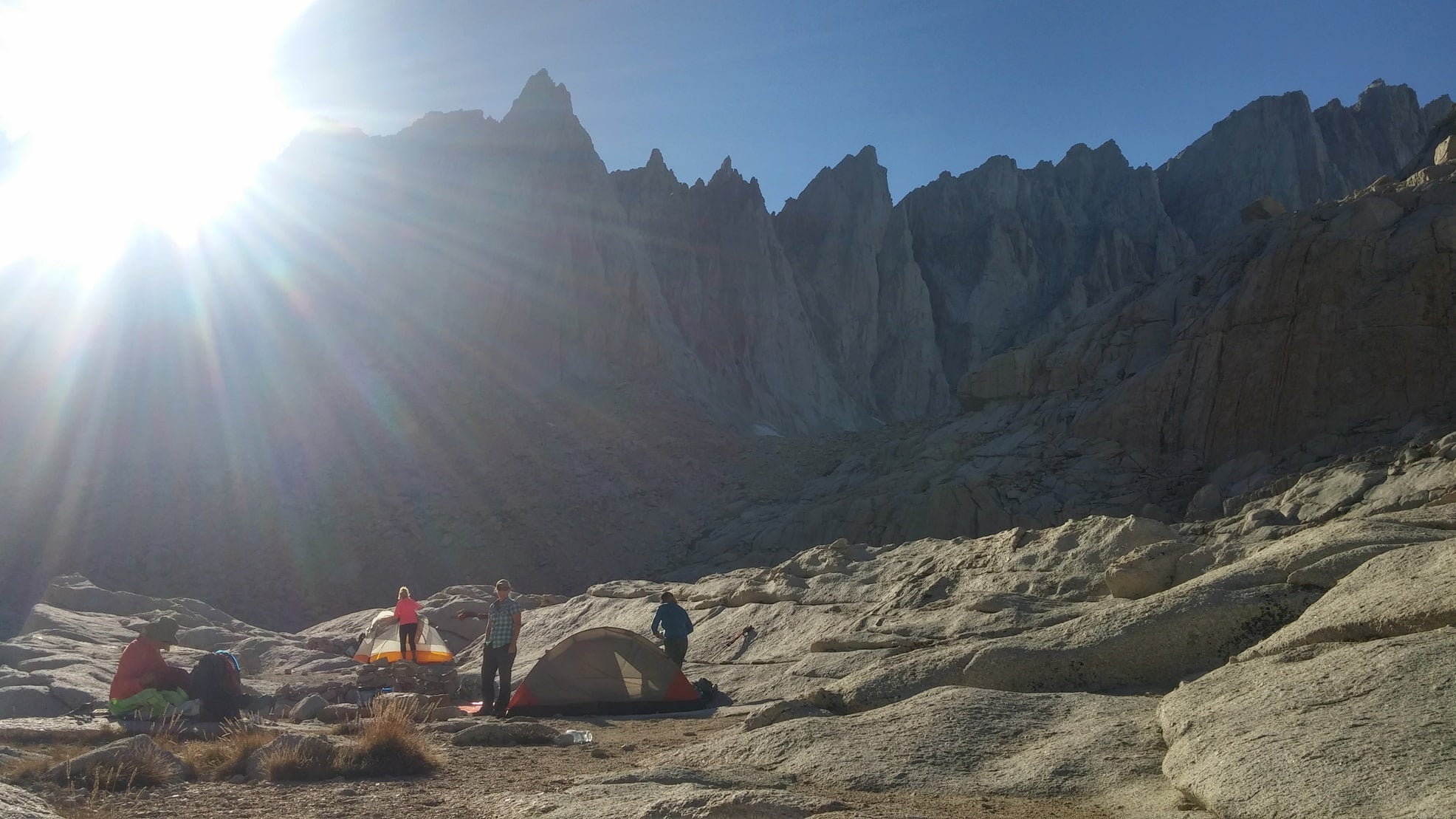 Shining sun peaking out from the jagged Sierra range with cmapers hanging out around tents in the forgeground.