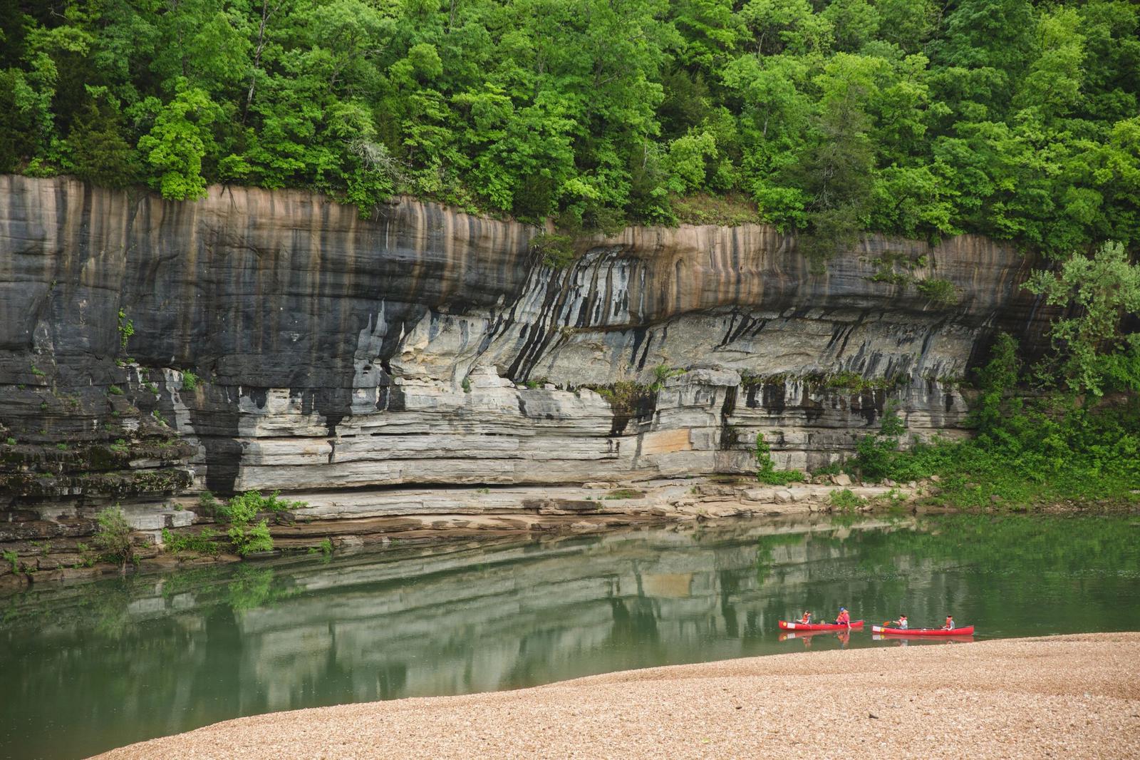 Two red canoes with people in them floating on a waterway past a large rock wall surrounded by green lush forest.
