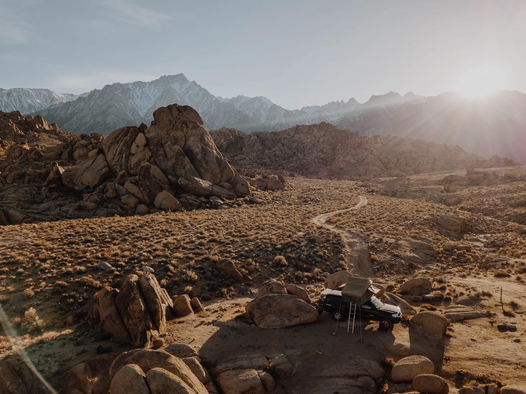 Sun shining over the jagged snowcapped sierras with a truck camper parked in the desert below at Alabama Hills.