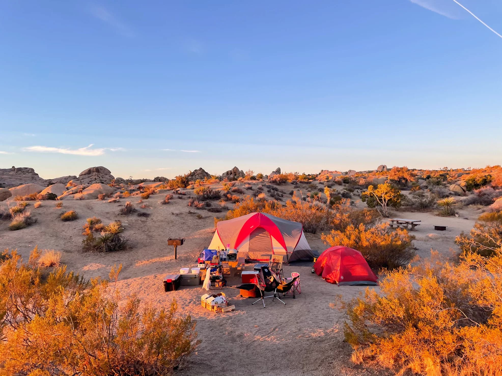 Golden hour at a desert campsite with a red tent surrounded by brush and red rock formations.