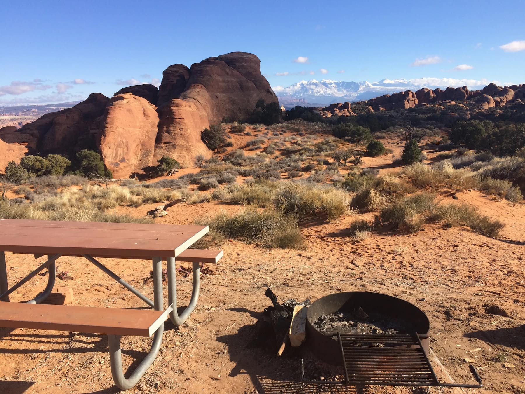 Campsite with picnic table and fire ring in red rocky desert with snow capped mountain landscape in the distance.