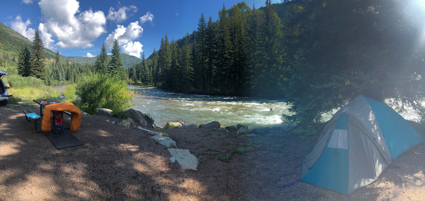 Panoramic view of campsite along flowing river with tent and picnic table.