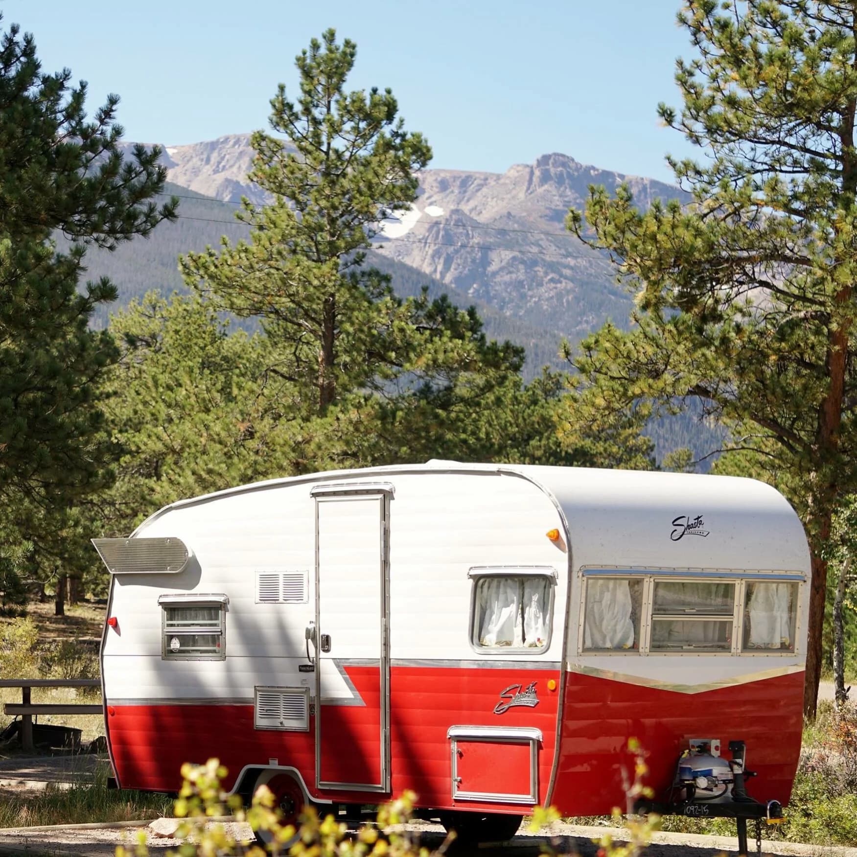 Vintage red and white trailer parked beside pine trees below mountains.