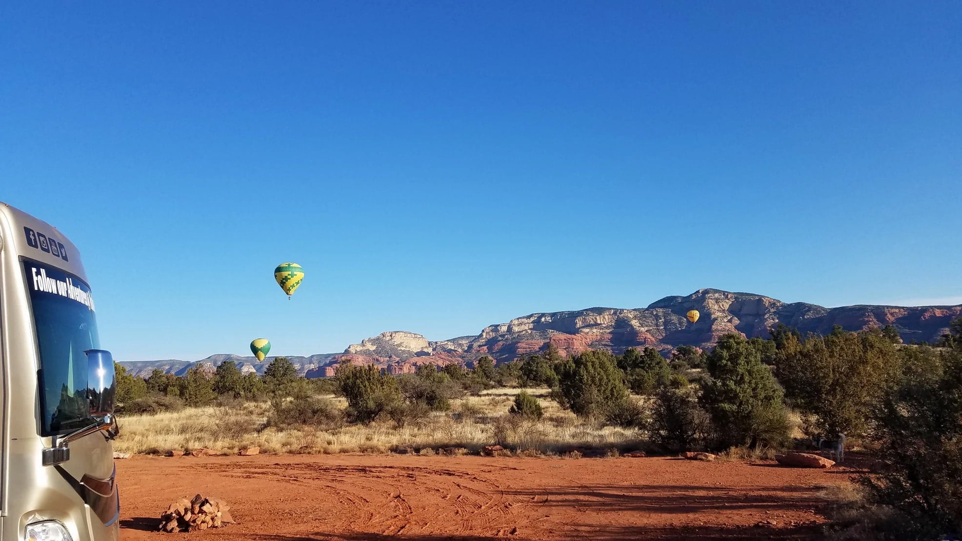 Hot air baloons rising above the desert above rocky mountains in the distance with an RV parked at a desert campsite in the foreground.