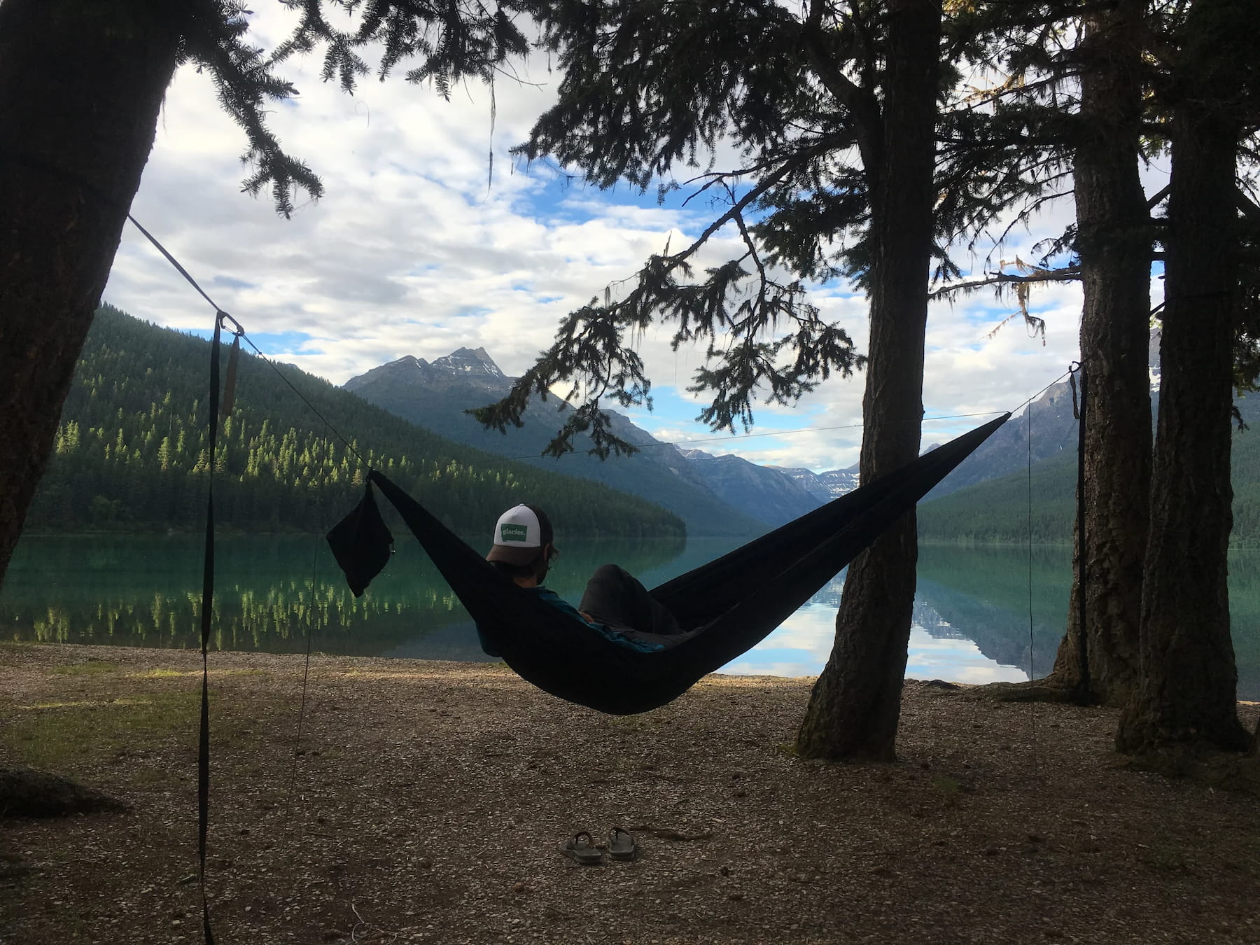 Person in backwards hat in hammock in the forest beside a lake with mountains in the distance.