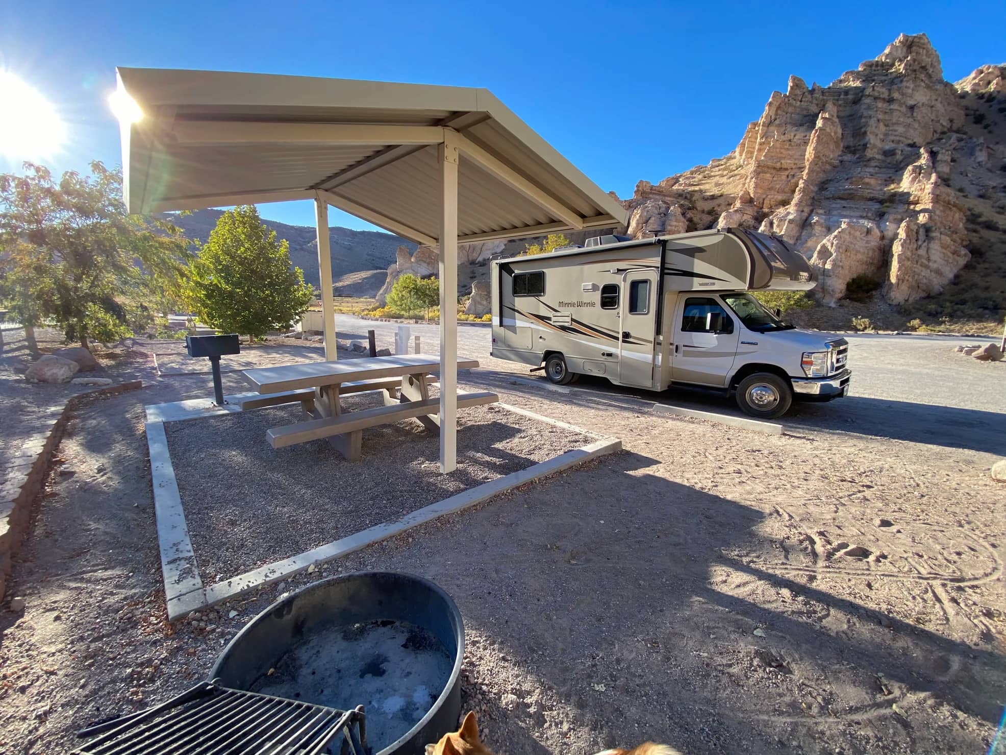 RV parked beside lean-to with picnic table and fire pit with jagged rocks in the distance.