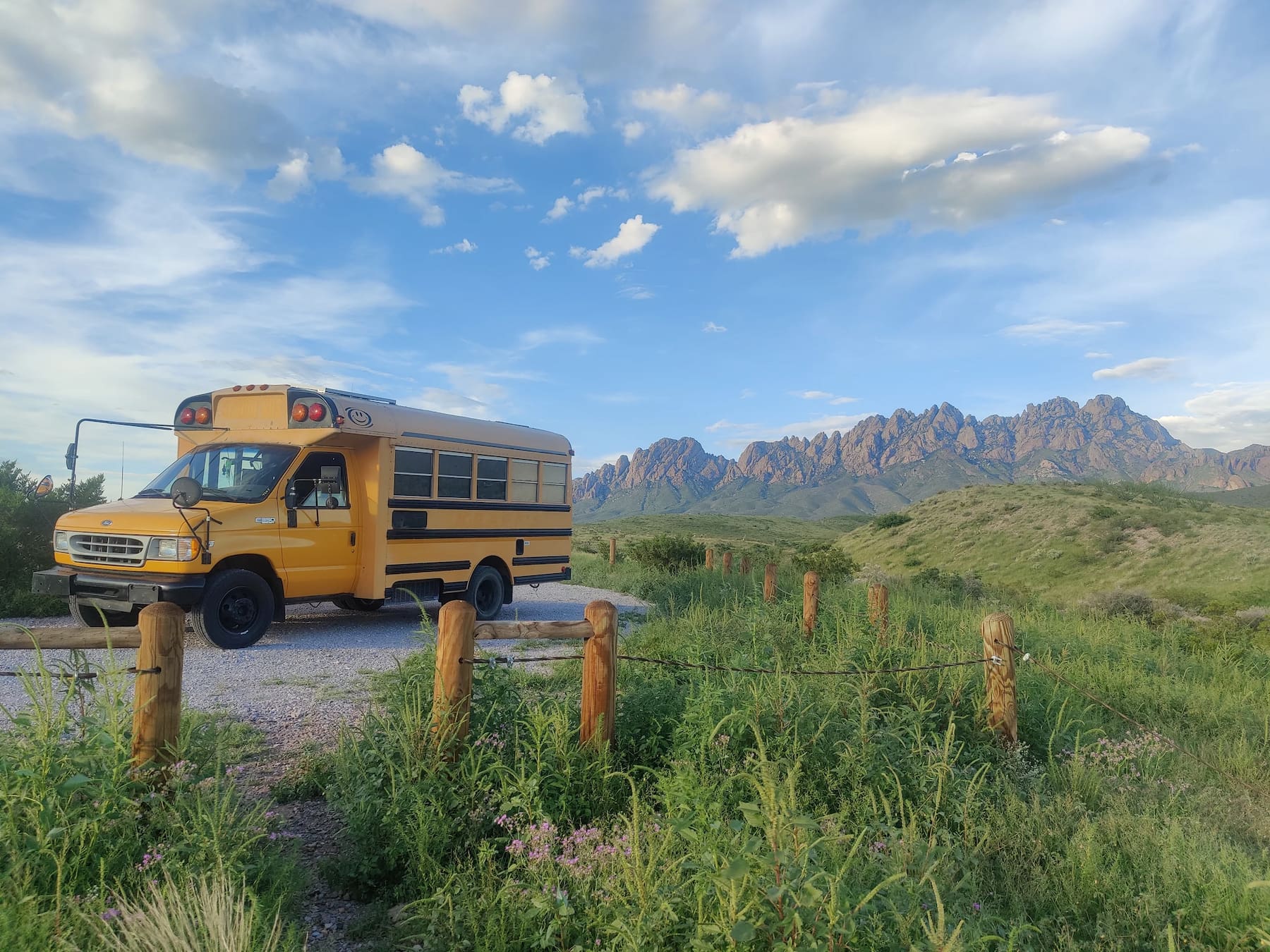 Short yellow school bus parked at a campsite beside wooden fence posts in a field and the Sierra mountains in the background.