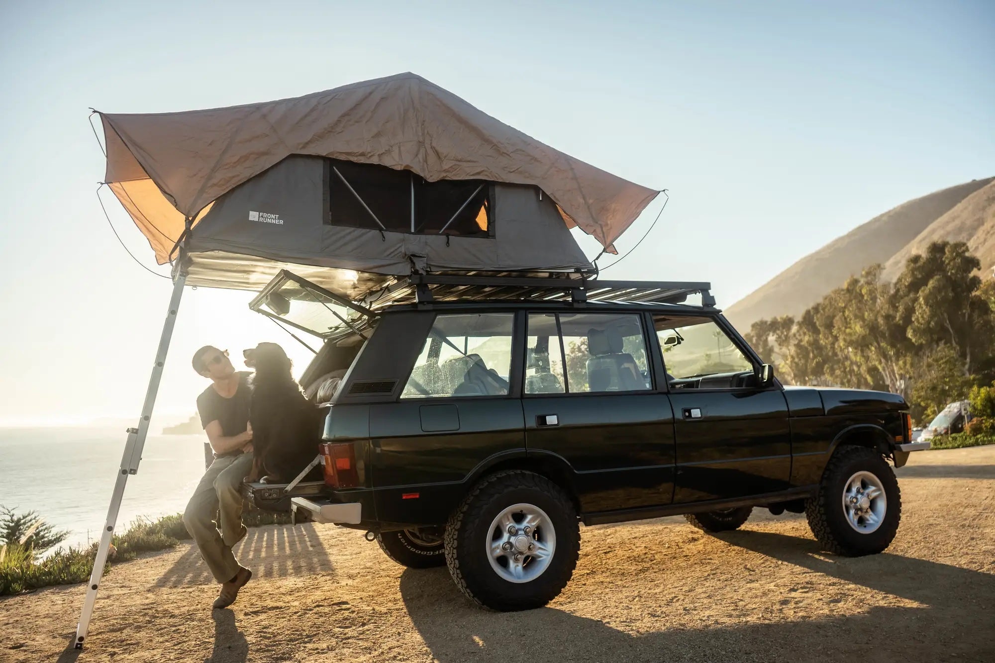 A guy and a dog sit on the tailgate of a car under a rooftop tent