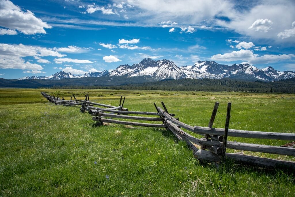 Wooden slit rail fence fades into the distance across a green and lush pasture toward the snow capped Sawtooth Mountain Range with a blue sky and white clouds above.